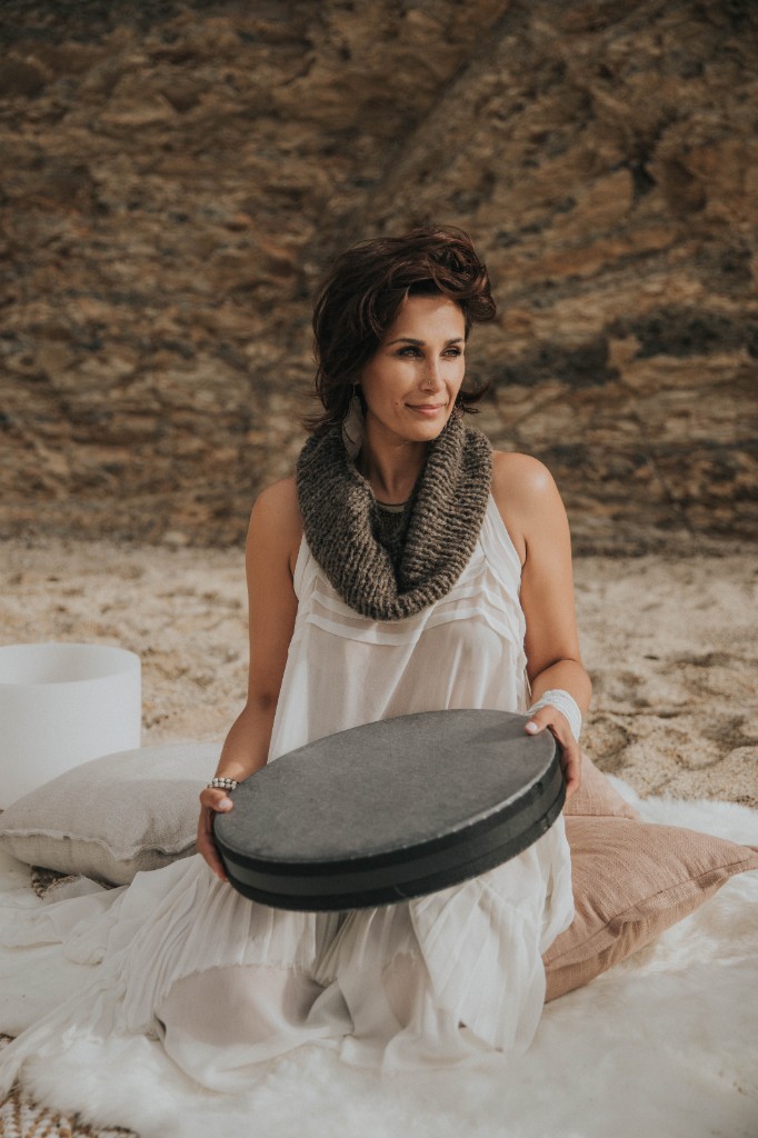 Tolo seated on the beach with a frame drum and crystal singing bowl